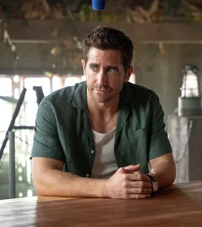 Two men sitting at a wooden table in a rustic indoor setting, both with serious expressions and casual shirts.