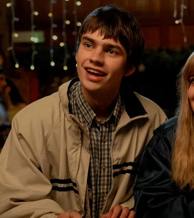 Teen boy and girl smiling and sitting together in a warmly lit indoor setting with people in the background