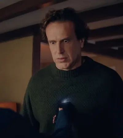 A man in a green sweater looks surprised as a woman with a ponytail holds out her hand indoors.