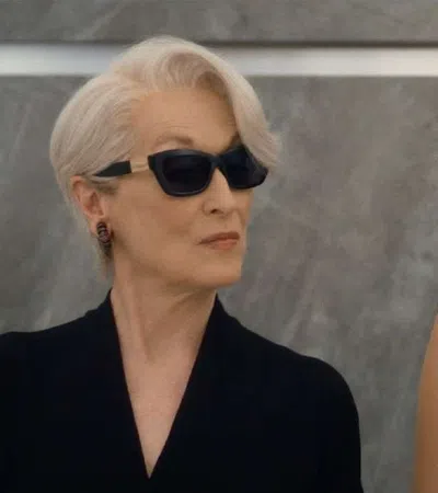 Two women in elegant attire, one with short white hair and sunglasses, the other with long dark hair and pearls.