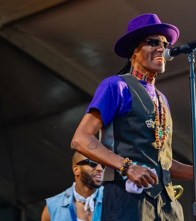 Musician in purple hat and vest singing into microphone while playing tambourine on stage at outdoor event.