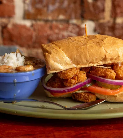 Crispy fried shrimp po' boy sandwich with lettuce, tomato, onion, served with a side of rice and beans in a blue bowl.