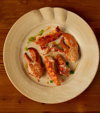 Grilled lobster claws and tails served with colorful sauce drizzles on a beige ceramic plate over a wooden table.