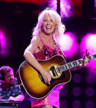 Female country singer in pink dress playing acoustic guitar on stage with vibrant purple lighting and smiling joyfully.