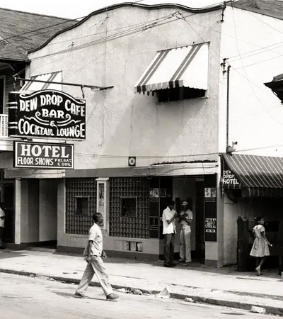 Black and white street scene with Dew Drop Cafe bar, cocktail lounge, hotel, and people walking and standing outside.