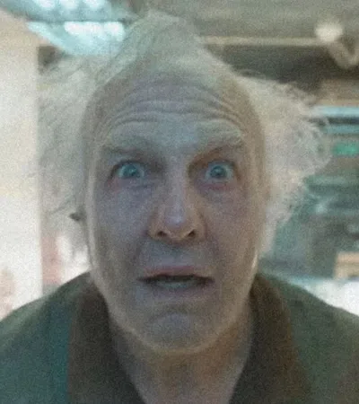 Older man with wild white hair and surprised expression looking directly at the camera indoors.