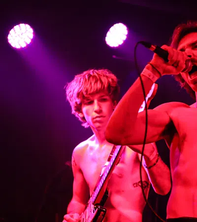 Two shirtless male musicians perform on stage under pink and purple lights, one singing and one playing guitar.