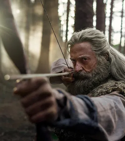 Focused bearded man with long hair aiming a bow and arrow in a misty forest during daytime