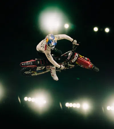 Motocross rider performing a stunt mid-air against bright stadium lights in a dark arena.