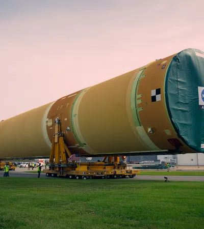 Large NASA Artemis rocket stage being transported on wheeled platforms outside a space facility at dusk.