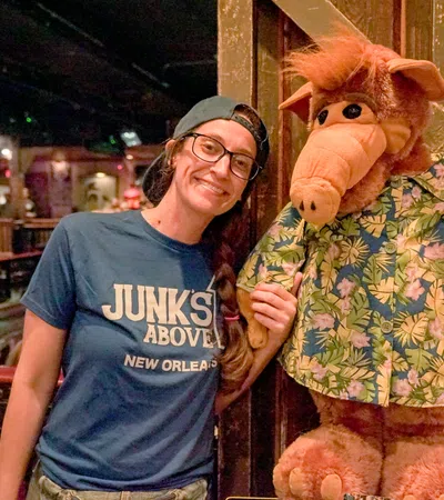 Smiling woman wearing glasses and a cap poses with ALF character doll in a tropical shirt inside a dimly lit venue.