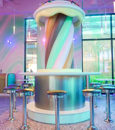 Colorful twisted soft serve sculpture with a cherry top in a retro diner booth surrounded by chrome stools.
