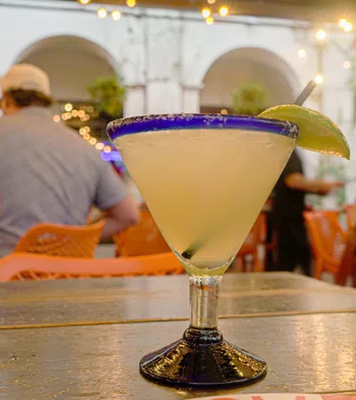 Margarita cocktail with a lime wedge on a wooden table in a lively restaurant setting with people and string lights.