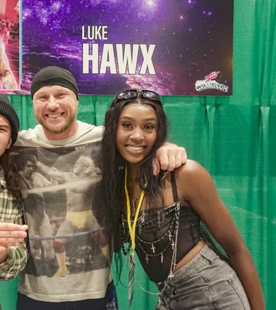 Three people smiling and posing in front of a banner featuring wrestler Luke Hawx at a convention booth.