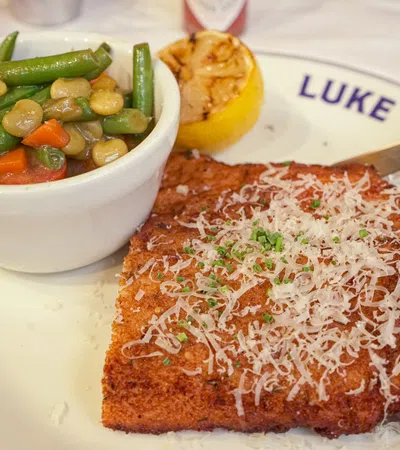 Breaded meat cutlet topped with grated cheese and herbs served with mixed vegetables and grilled lemon on a white plate.