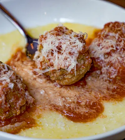 Three meatballs topped with grated cheese served in marinara sauce over creamy polenta in a white bowl.