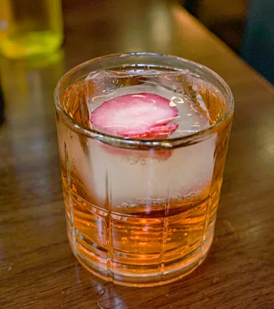 Glass of amber cocktail with large ice cube and radish slice on wooden table in a bar setting
