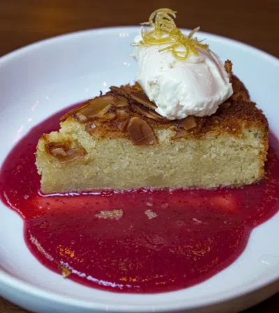 Slice of almond cake with whipped cream and lemon zest on raspberry sauce in white bowl.