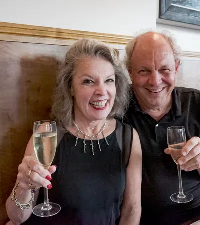 Smiling older couple in black outfits holding champagne glasses celebrating indoors at a restaurant.