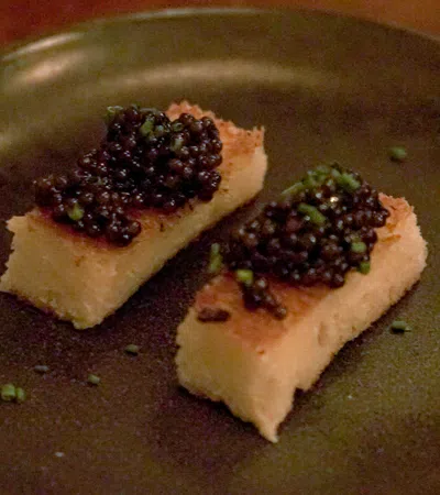 Two rectangular pieces of toasted bread topped with black caviar and garnished with chopped chives on a dark plate