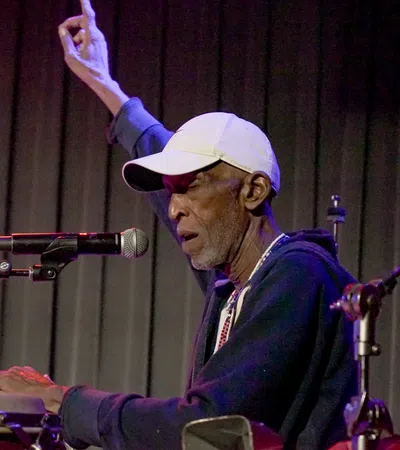 Elderly male musician playing congas and singing into a microphone with one hand raised on stage.