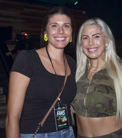 Two smiling women posing indoors, one in black top and the other in camouflage crop top with long blonde hair.