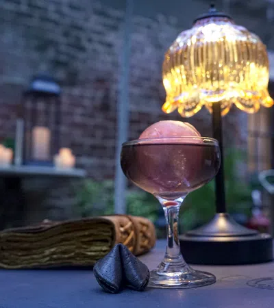 Elegant cocktail glass with a pink spherical garnish on a table beside a decorative lamp and fortune cookies.
