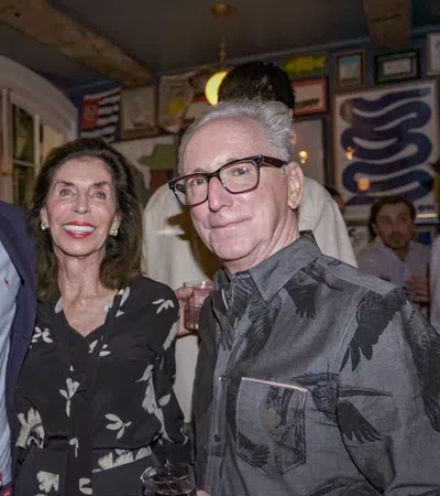 Three adults smiling together indoors, dressed in semi-formal attire at a social gathering with framed art on walls.