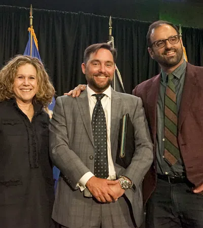 Three professionally dressed people smiling with flags in the background at an indoor event.