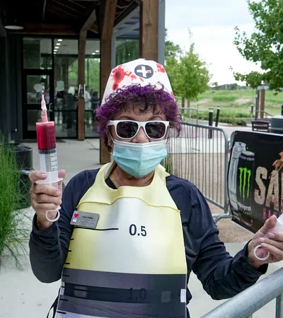 Person in nurse costume wearing sunglasses and mask holding two large syringes outside a building.