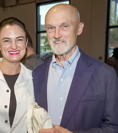 Smiling woman and elderly man standing indoors at a social event with large windows in the background.