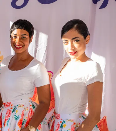 Three smiling women wearing white tops and floral skirts stand in front of a colorful backdrop with plants and coral designs.