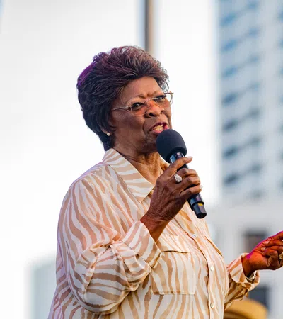 Elderly woman in striped blouse speaking into microphone at outdoor event with city building background