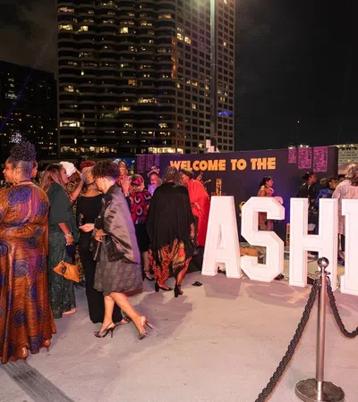 People dressed in elegant attire gather at a nighttime outdoor event with large illuminated ASHE letters and city buildings in the background