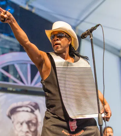 Musician wearing a white cowboy hat and playing a washboard while performing on stage with a microphone.