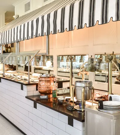 Bright buffet line with white tiles, striped canopy, copper pots, plates, and food warmers in a dining area.