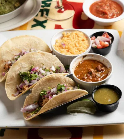 Three tacos with chopped onions, cilantro, and meat served with rice, beans, salsa, lime, and green sauce on a white plate