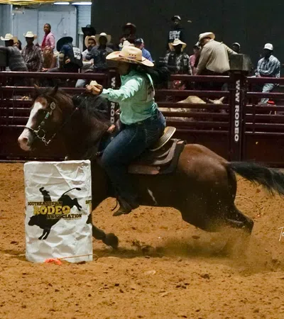 Cowgirl riding horse fast around barrel during rodeo competition in indoor arena with spectators