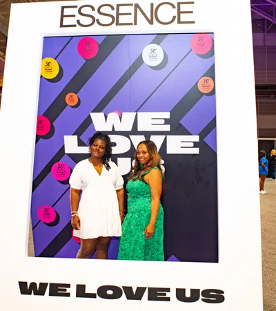 Two women posing inside a large Essence-themed photo frame with the text We Love Us at an indoor event.