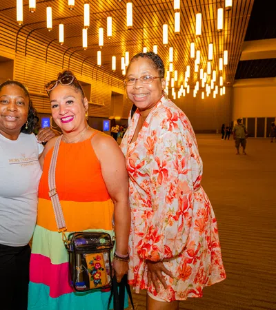 Three women smiling indoors under modern ceiling lights with wooden wall panels in a spacious venue.