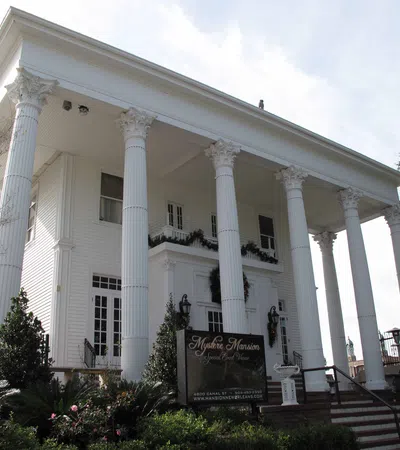 White Greek Revival mansion with tall Corinthian columns and manicured garden in front under a clear sky.