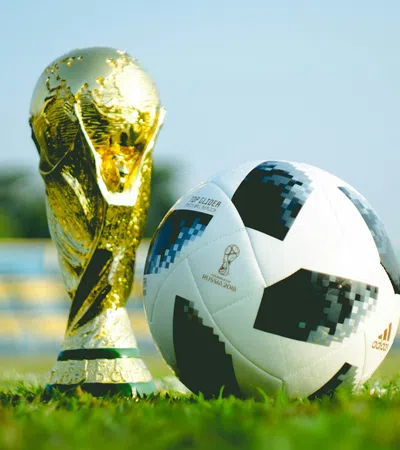 Golden FIFA World Cup trophy and official Adidas soccer ball on grass pitch with blurred stadium seats background.