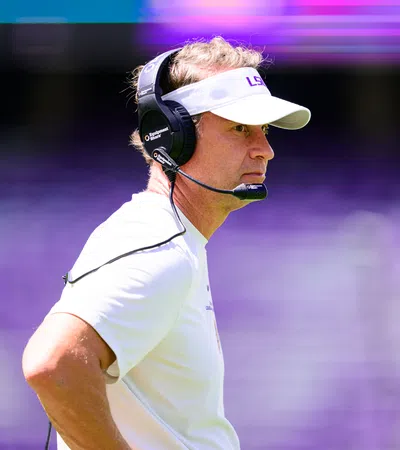 Football coach wearing LSU visor and headset focused during a game with blurred purple stadium background