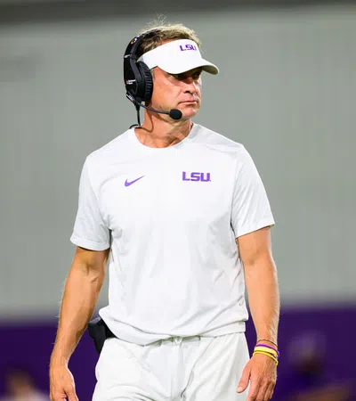 LSU football coach in white LSU gear and visor wears a headset on the sidelines during a game.