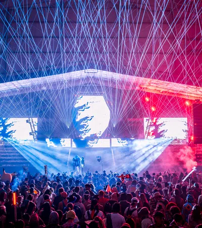 Crowd enjoying a nighttime concert with vibrant laser lights and colorful stage effects under a large canopy.