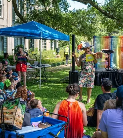 Woman reads a children's book outdoors to a seated diverse group of kids and adults on a sunny day