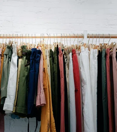 Variety of colorful pants and trousers hanging on a wooden rack against a white brick wall.