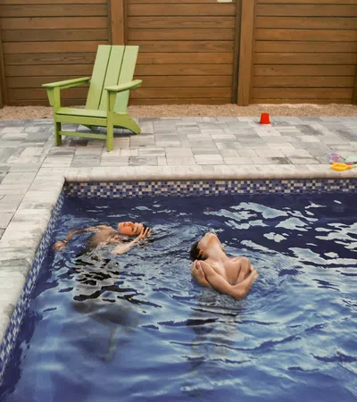 Two boys swimming and playing in a backyard pool next to a green Adirondack chair on a stone patio.