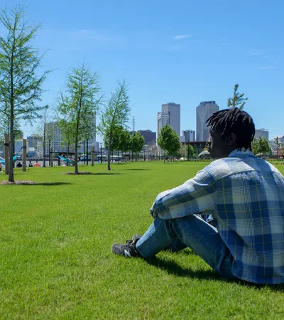 Young man in plaid shirt sitting on green grass with city skyline and trees in background on clear day
