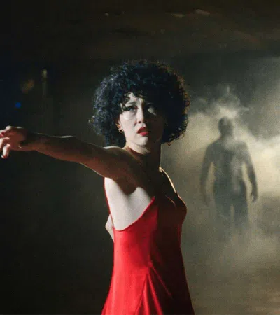 Woman in a red dress reaching out hand in a dark room with mist and a shadowy figure in the background
